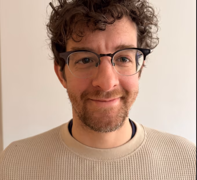 Smiling man with curly dark hair, beard, glasses, and beige sweater against a plain background.