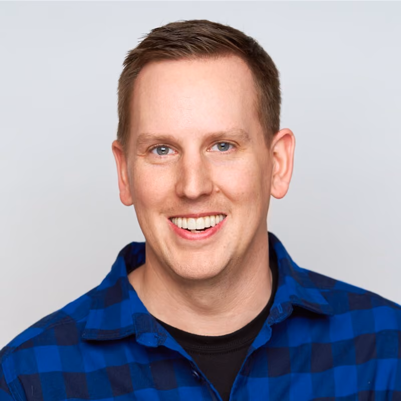 Smiling man with short brown hair wearing a blue checkered shirt and black undershirt on light background.
