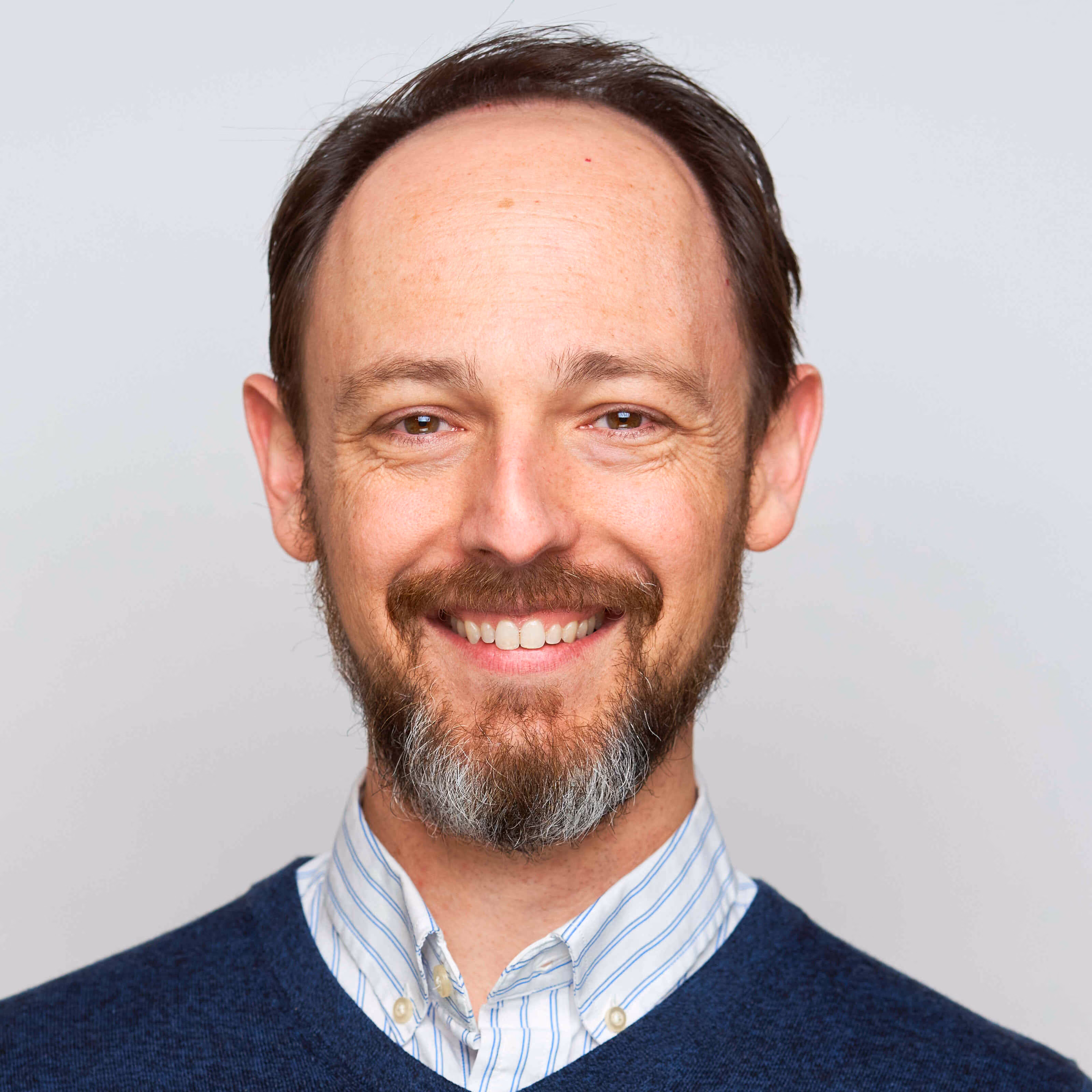 Smiling man with beard and mustache wearing a striped shirt and blue sweater against a gray background.