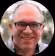 Smiling man wearing glasses outdoors with trees and a building blurred in the background.
