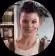 Smiling woman with short hair and a necklace, indoors with blurred bookshelves in background.