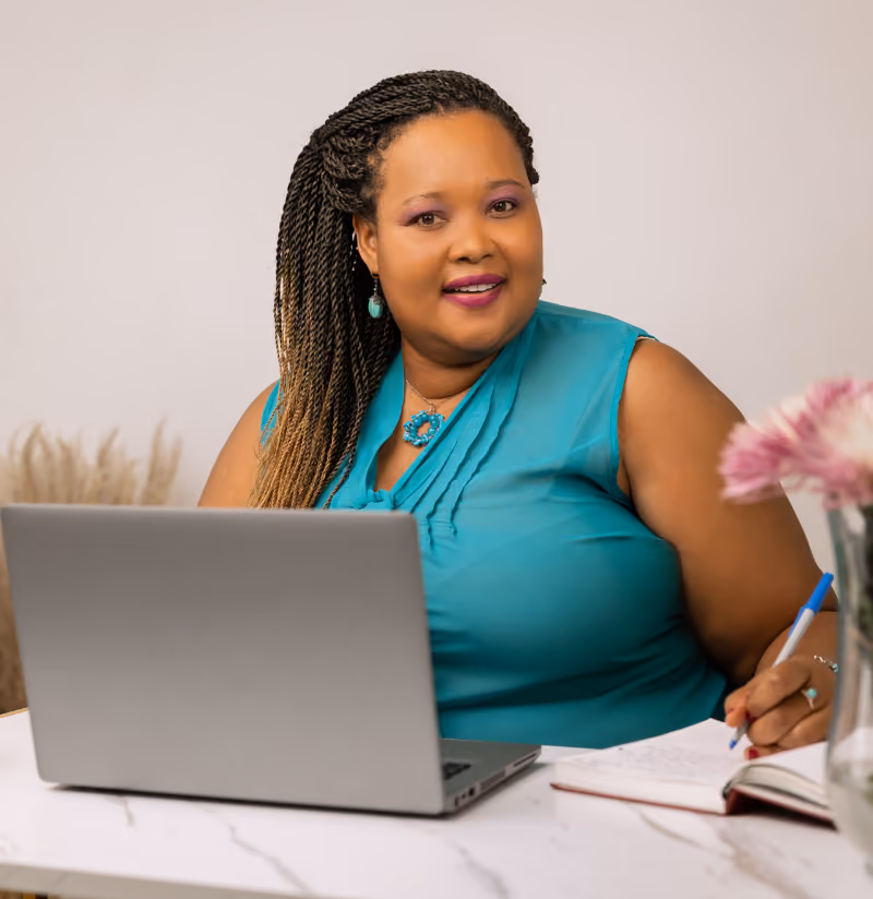 Woman with braided hair in turquoise top smiling at desk with laptop and notebook.
