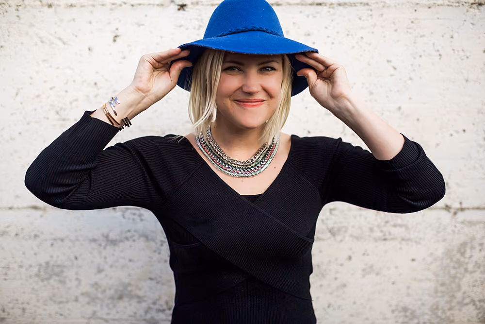 Smiling woman wearing a blue hat, black top, and layered necklaces poses against a textured wall.
