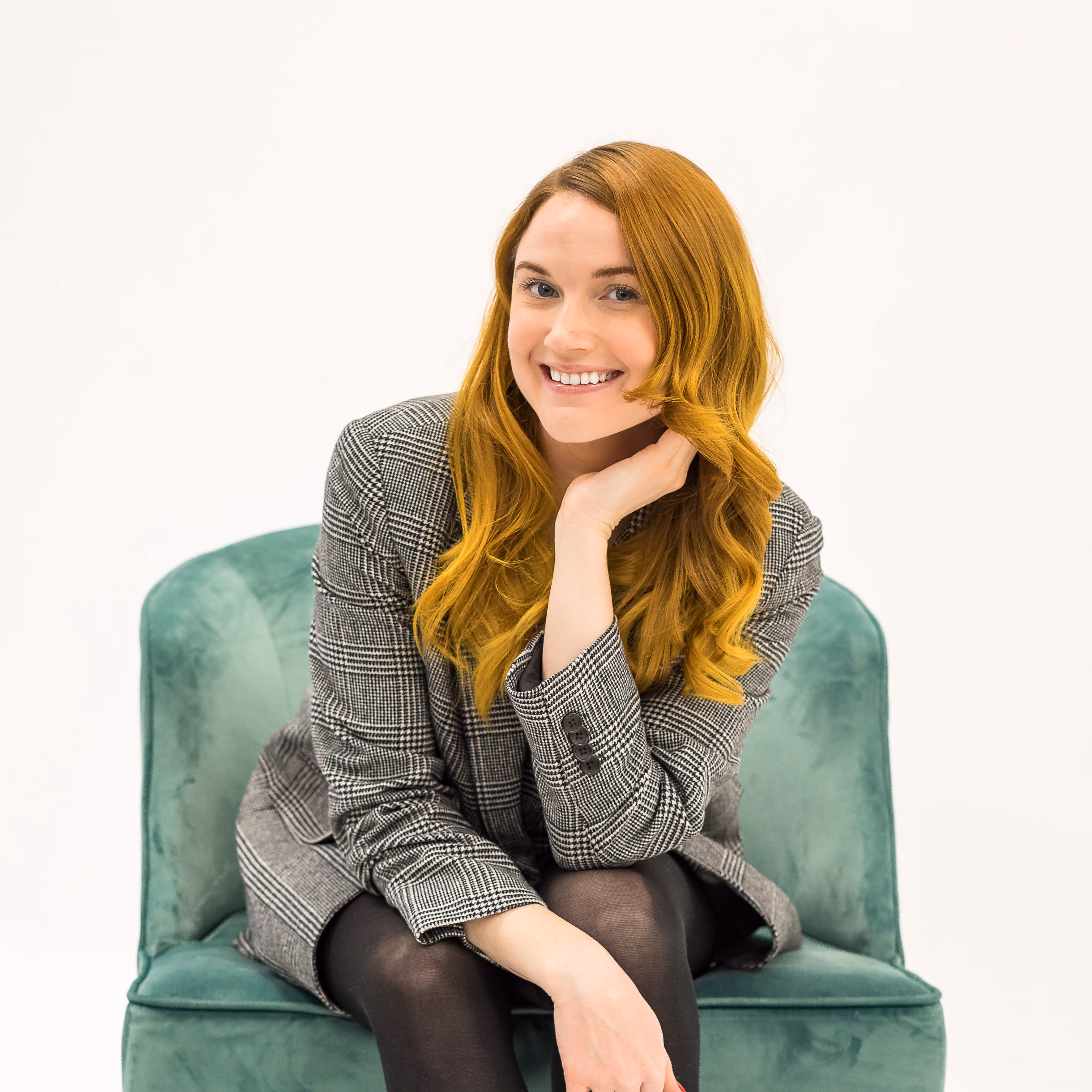 Smiling woman with long red hair wearing a gray plaid blazer sitting on a green chair.