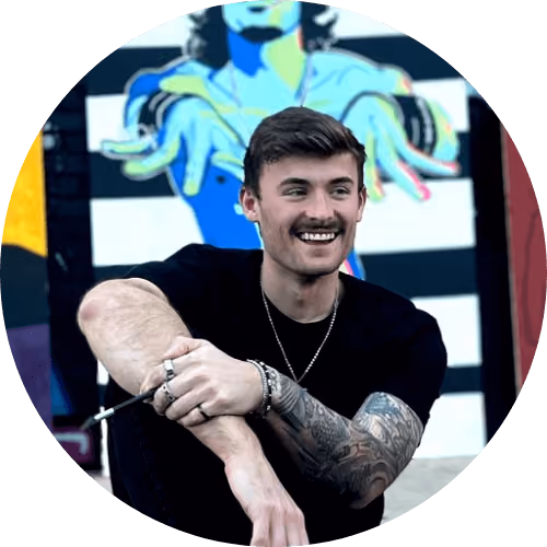 Smiling young man with dark hair and tattoos on arm sitting in front of colorful mural.