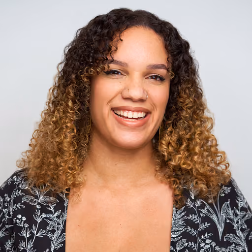Smiling woman with curly ombre hair wearing a black floral top against a plain background.