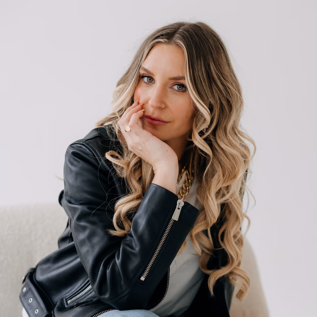 Woman with long curly blonde hair wearing a black leather jacket and gold chain necklace resting her head on hand.