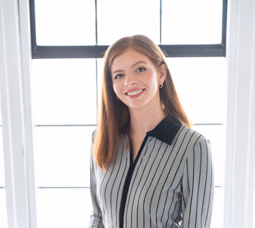 Smiling young woman with long red hair wearing a striped blouse standing in front of a window.