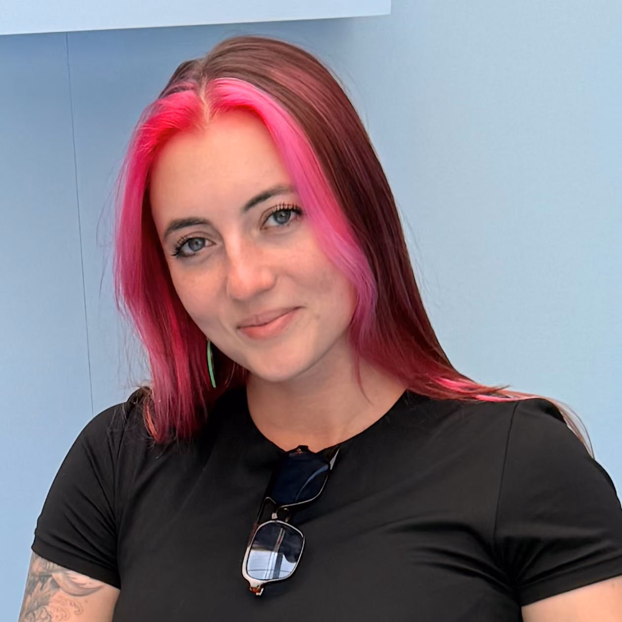 Woman with pink and brown hair, wearing a black shirt and sunglasses, smiling against a blue wall.