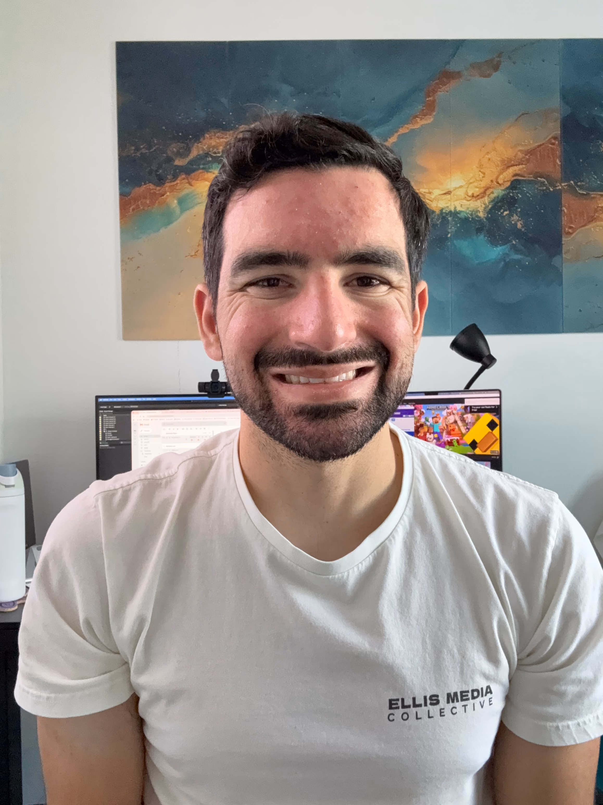 Smiling man with dark hair and beard wearing a white Ellis Media Collective t-shirt at a desk.