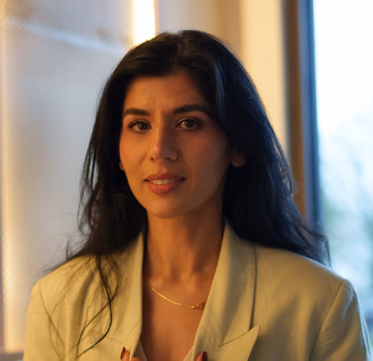 Woman with long dark hair wearing a beige blazer and gold necklace, smiling softly indoors by a window.