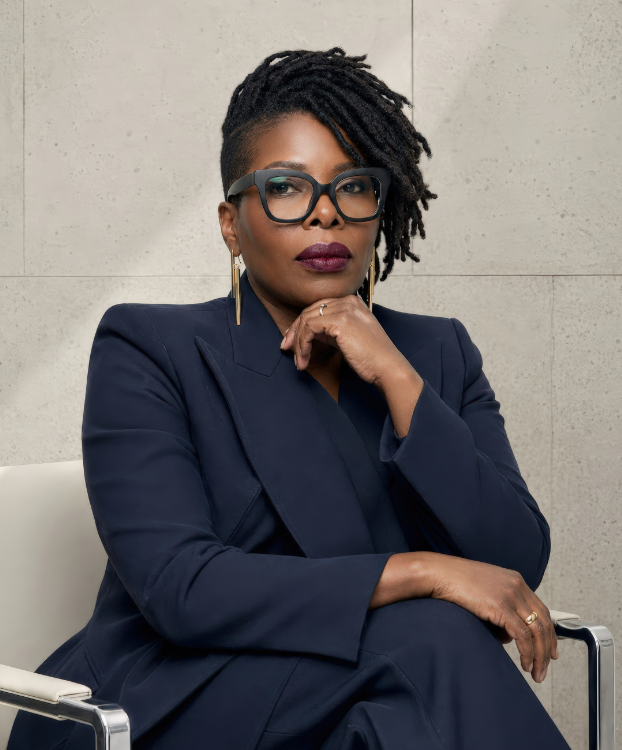Confident woman with black dreadlocks, glasses, dark lipstick, and navy blazer sitting thoughtfully.