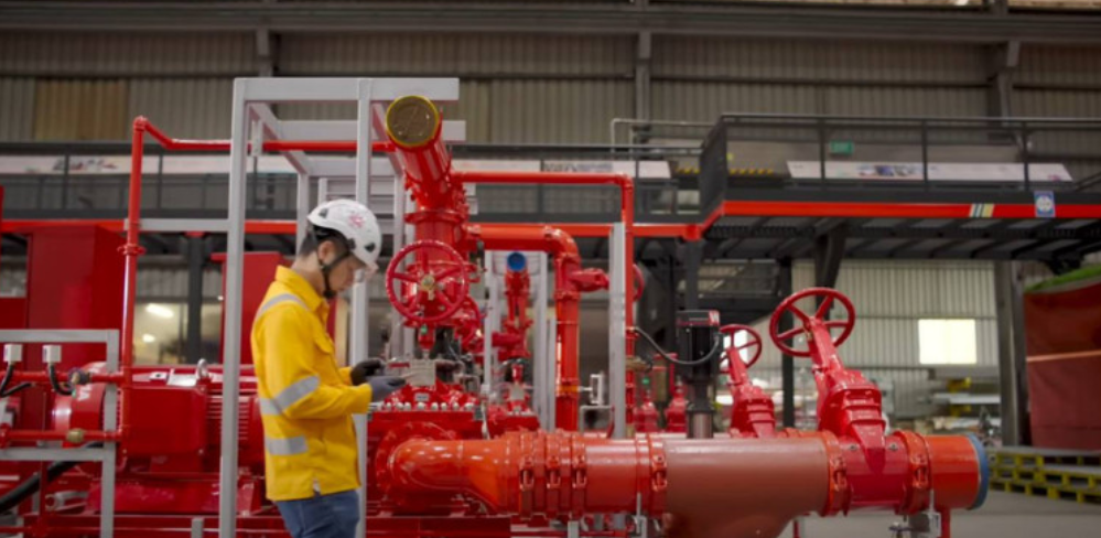 Worker in a yellow safety jacket and white helmet inspecting controls on red industrial piping in a factory setting.
