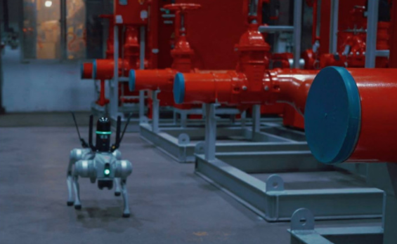 Small quadruped robot with antennas walking on a floor near large red industrial pipes.