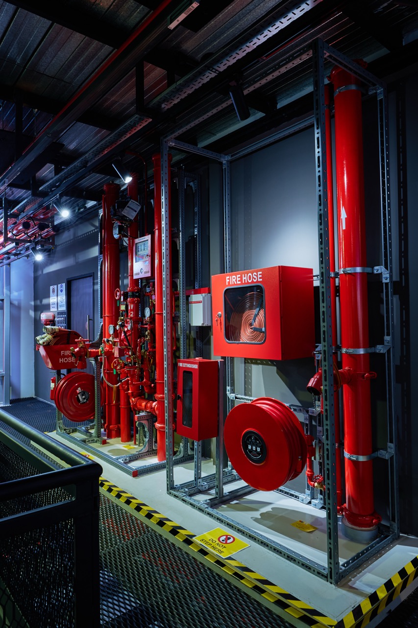 Industrial fire safety equipment installation with red fire hoses, pipes, and extinguishers along a wall in a maintenance area.