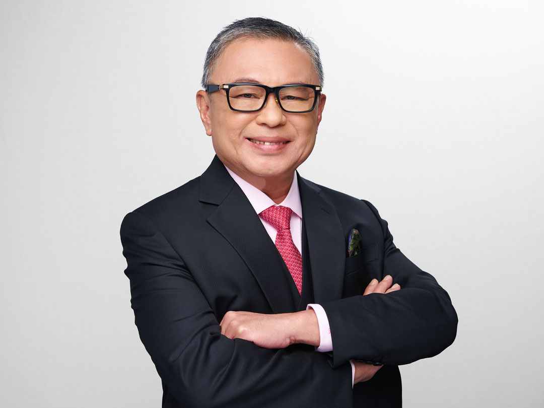 Smiling Dominic Ang in black suit and glasses with arms crossed against a plain background.