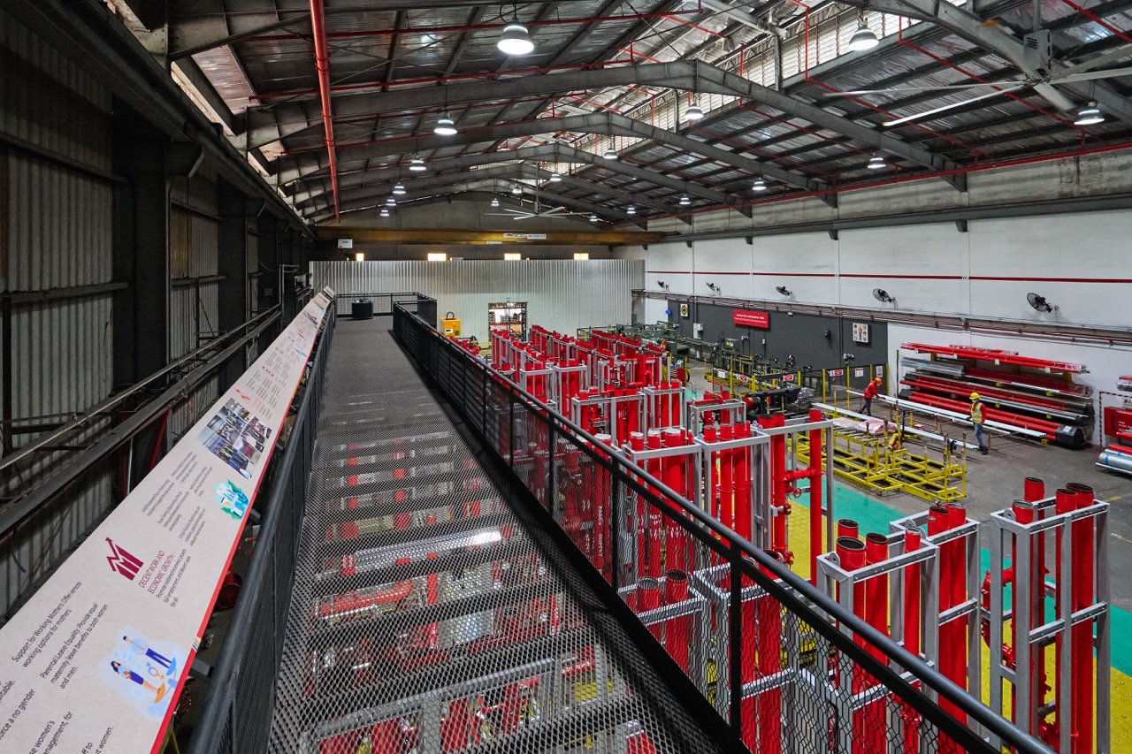 Interior of an industrial warehouse with red machinery, metal walkways, and two workers wearing helmets and safety vests.