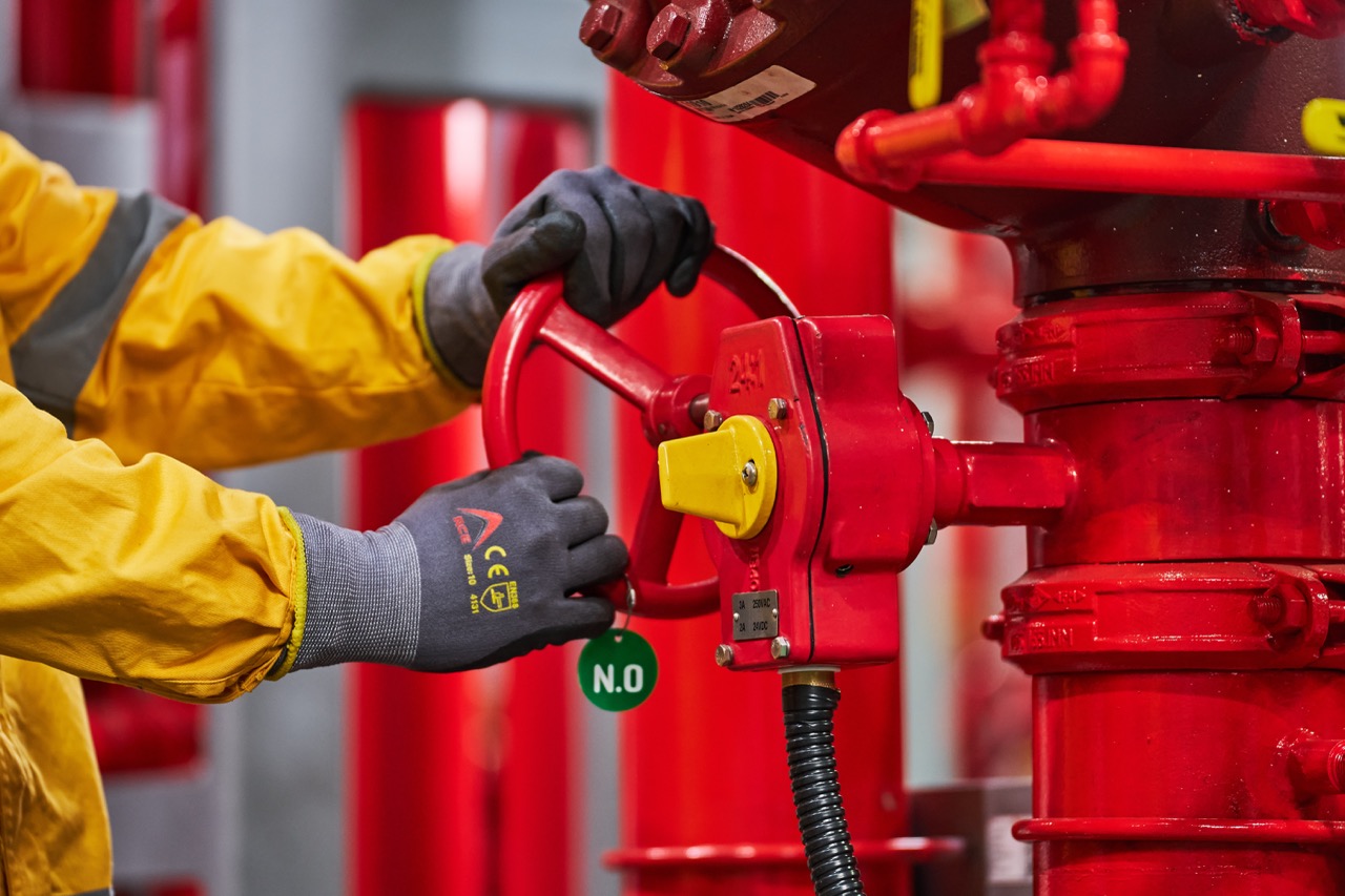 Person wearing yellow protective jacket and gray gloves operating a red fire hydrant valve inside a fire safety facility.