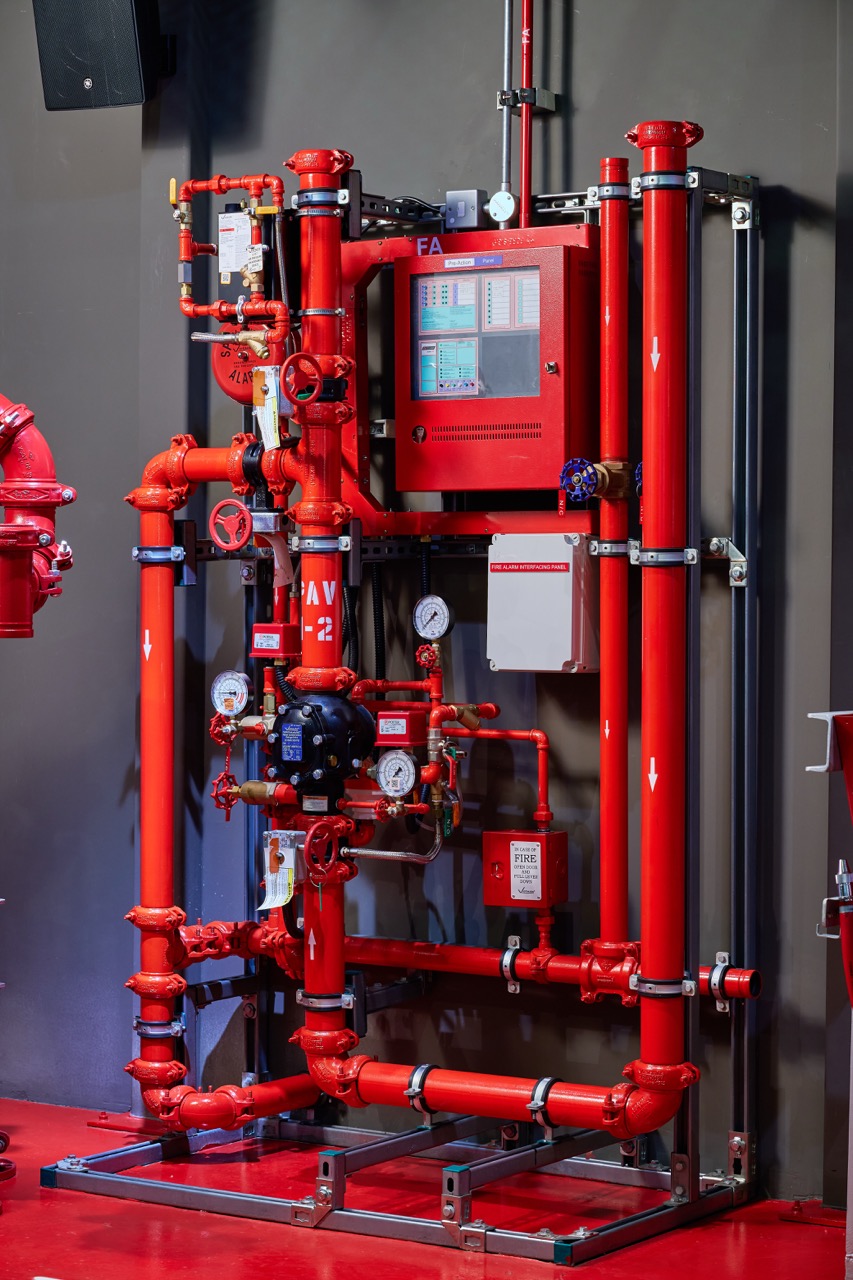 Red fire alarm control panel with connected red pipes, gauges, valves, and a fire alarm interfacing box mounted on a gray wall.
