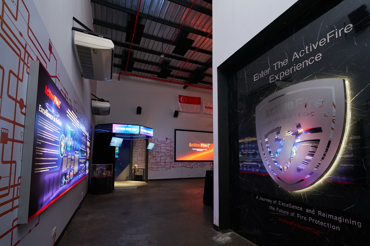 Interior view of the Active Fire Experience exhibition space with illuminated signs, a Singapore flag, and information displays on fire protection.
