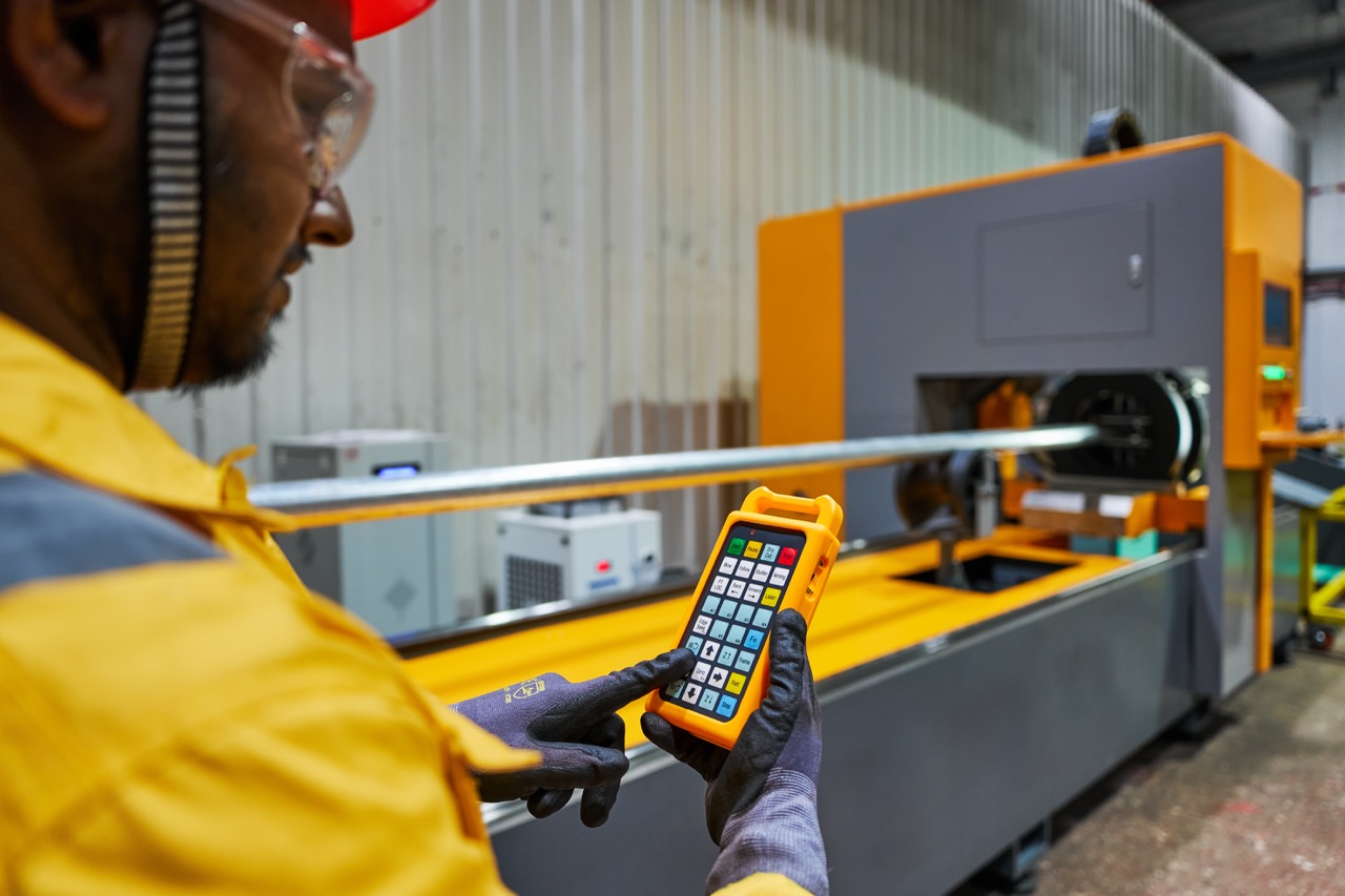 Factory worker in yellow safety gear and gloves operating industrial machine with handheld control panel.