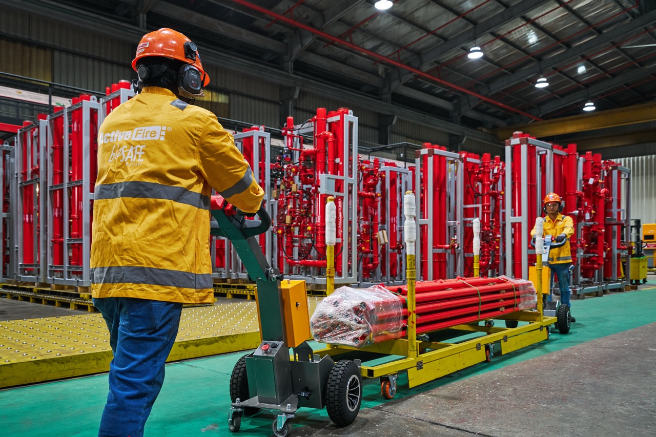 Two workers wearing yellow Active Fire jackets and helmets transporting red pipes on a yellow cart inside an industrial warehouse.