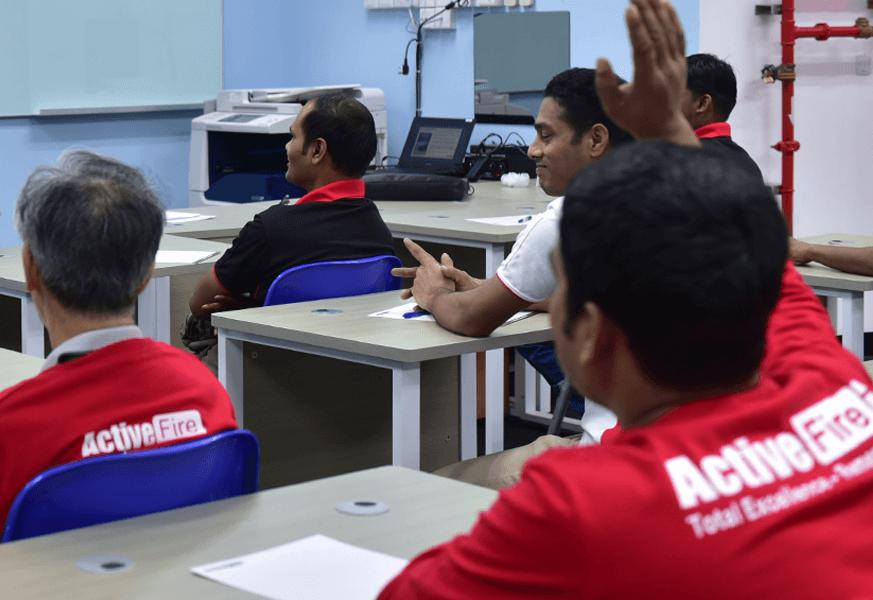Men seated at desks in a training room, one wearing a red shirt with 'Active Fire' text raising his hand.