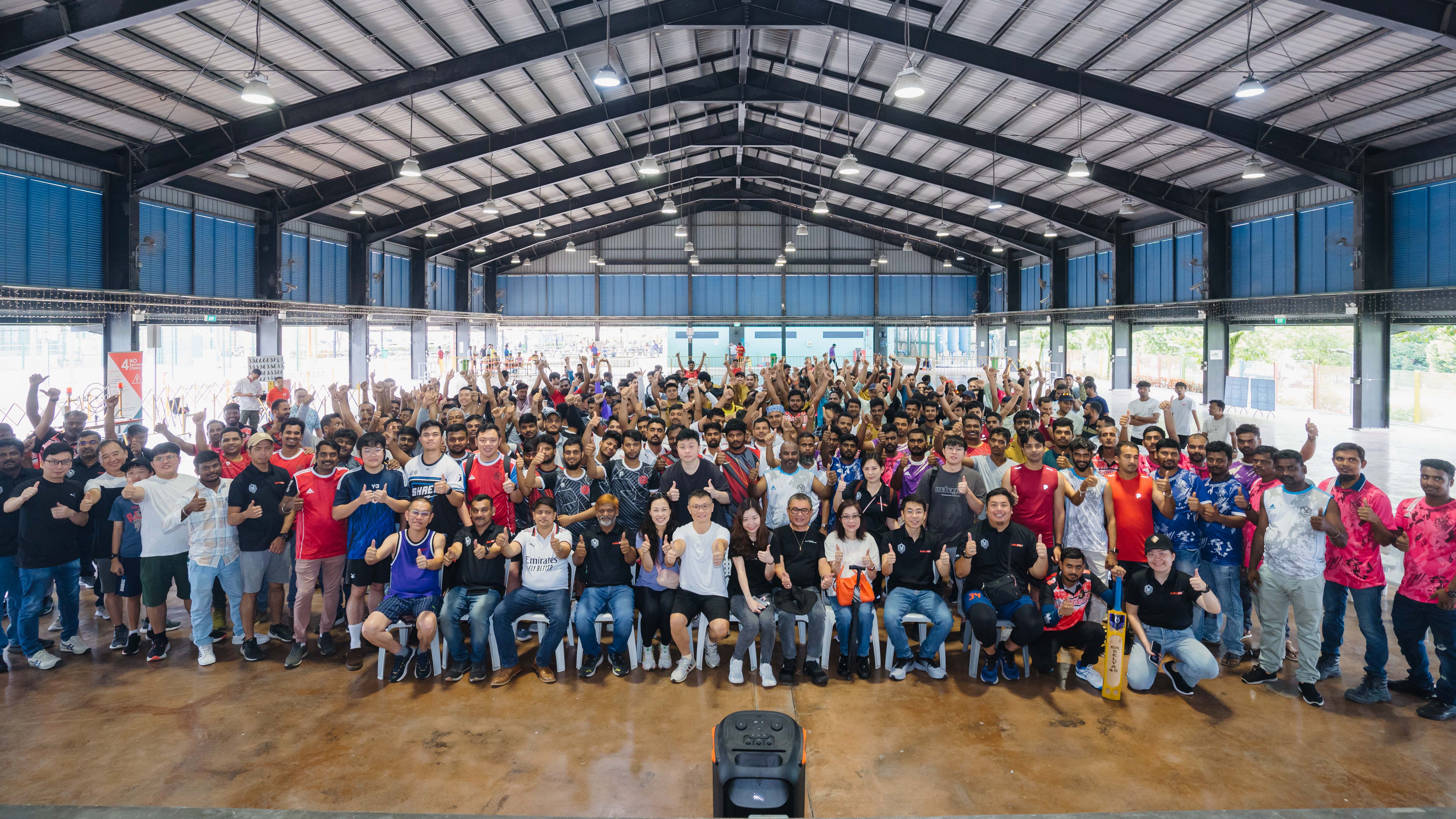Large group of diverse people posing inside a spacious industrial building, many giving thumbs-up gestures.