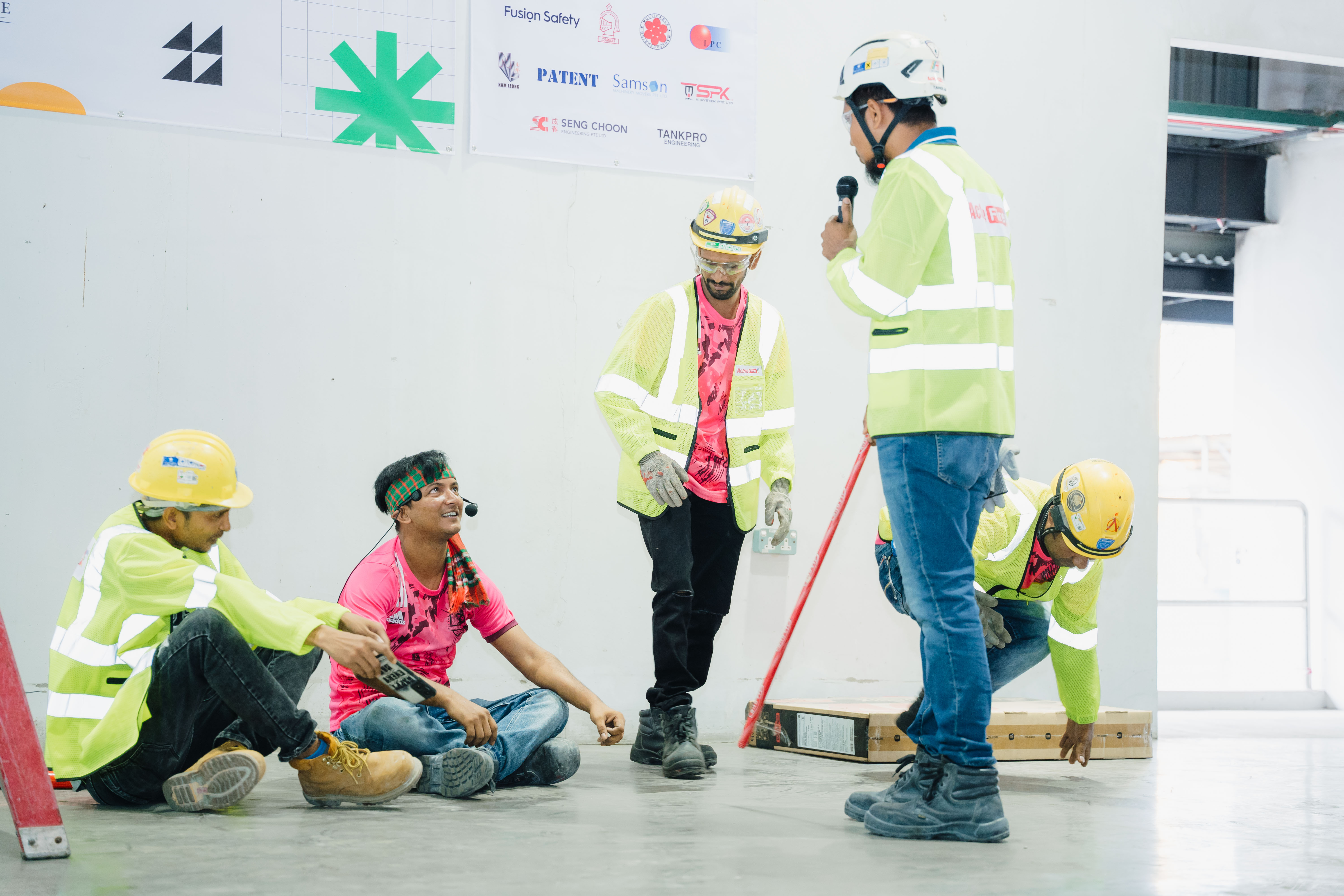 Five construction workers wearing helmets and safety vests engaged in discussion and tasks inside a building under construction.