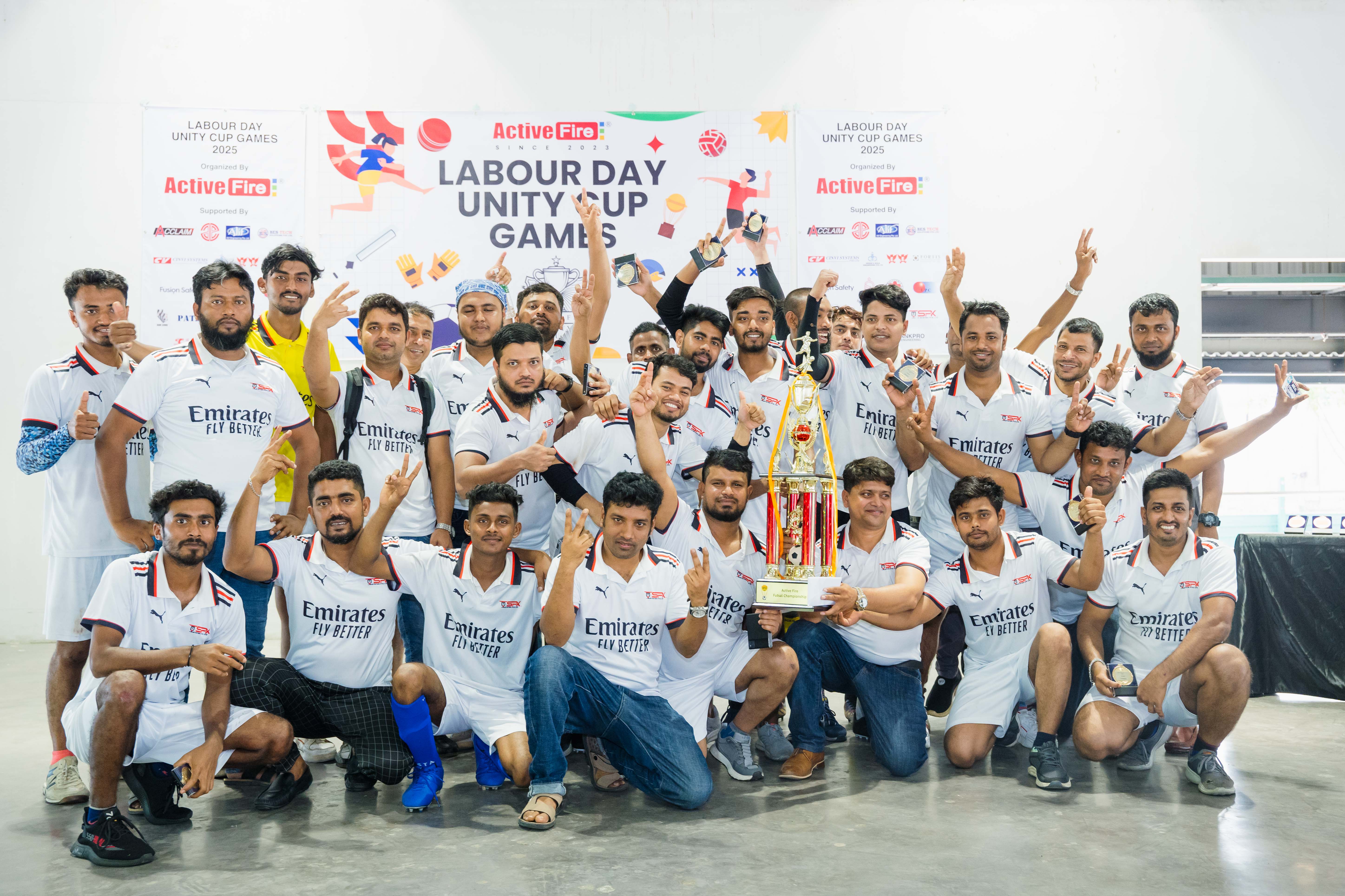 Group of men in white Emirates Fly Better sports jerseys posing with a large trophy and medals at the Labour Day Unity Cup Games event.