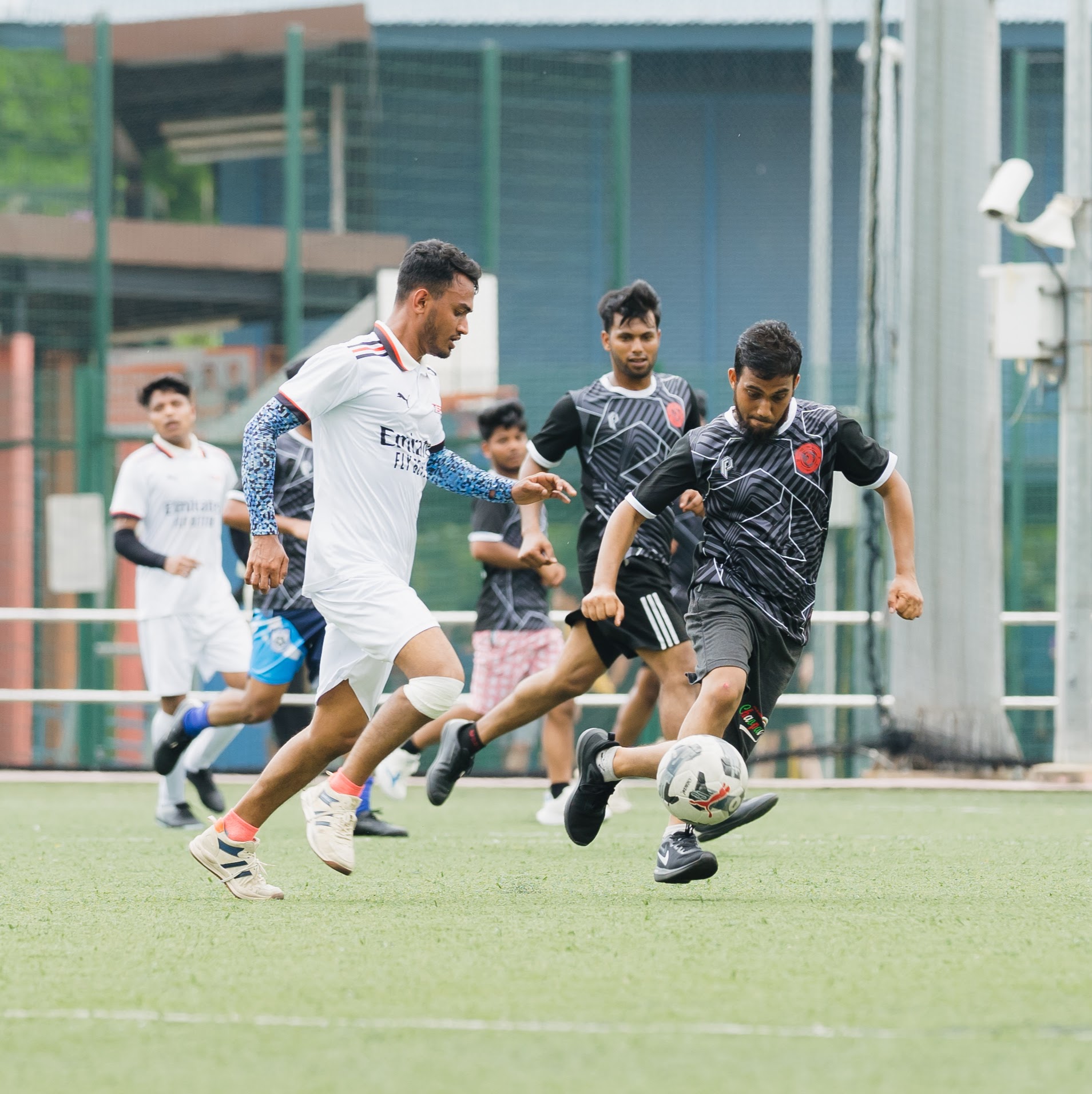 Men playing soccer on a green outdoor field, one in white kit and another kicking the ball in black kit.