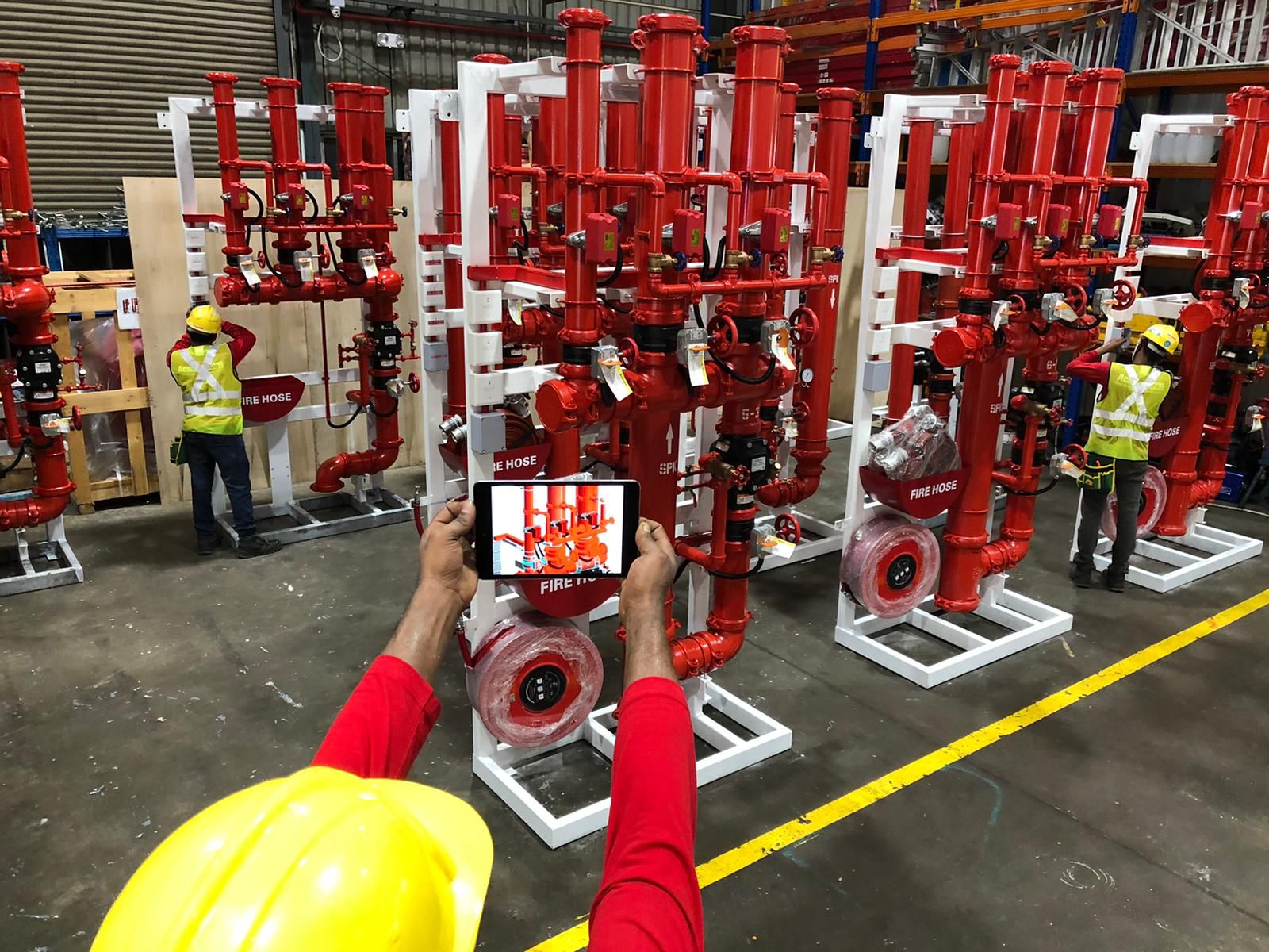 A person wearing a yellow hard hat and red shirt taking a photo of red fire hose equipment in a warehouse with two workers wearing yellow vests and hard hats in the background.