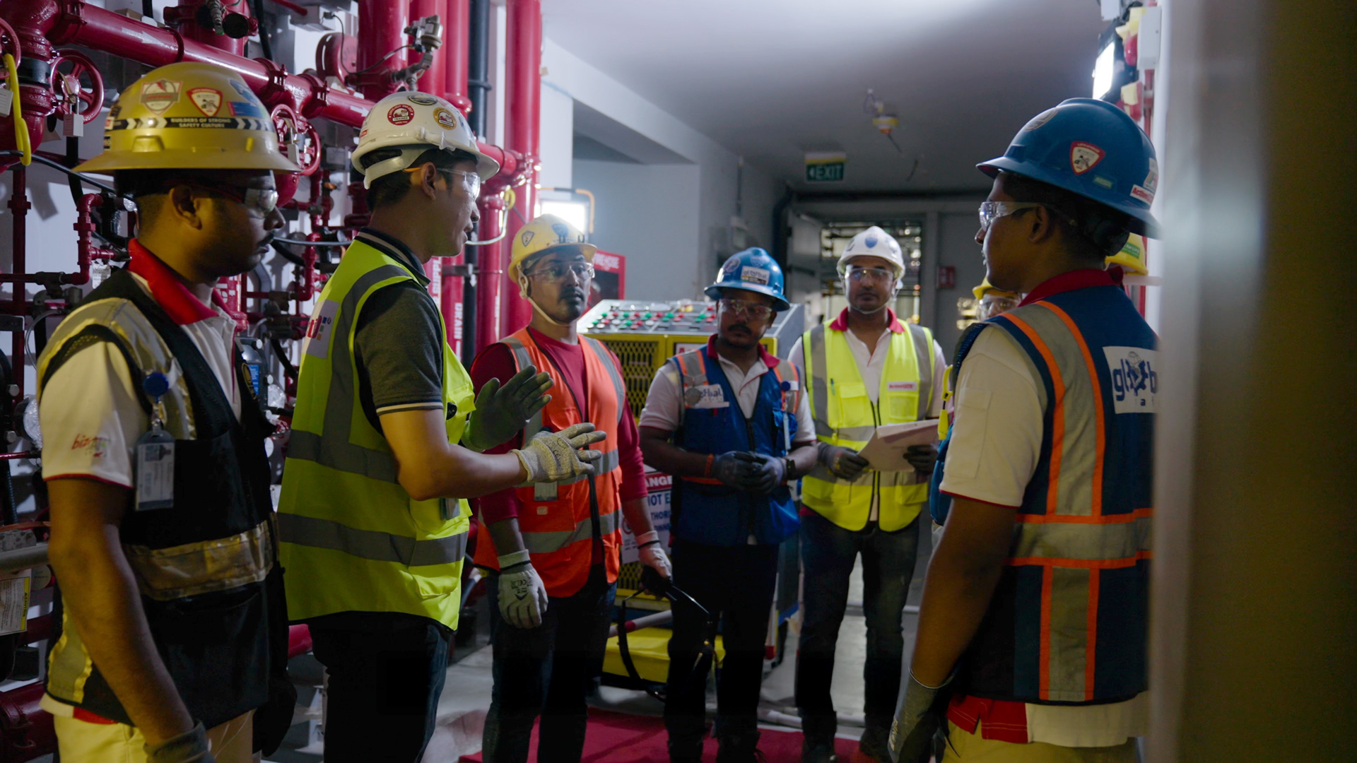 Group of seven construction workers in safety gear having a discussion indoors near red pipes.