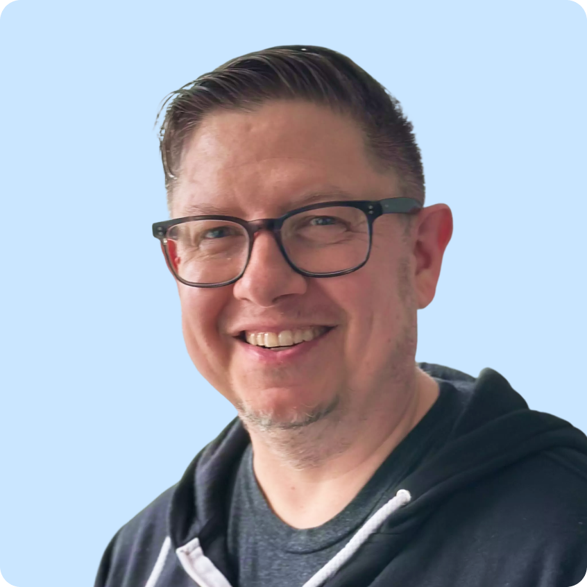 Smiling middle-aged man with short hair and glasses wearing a dark hoodie against a light purple background.