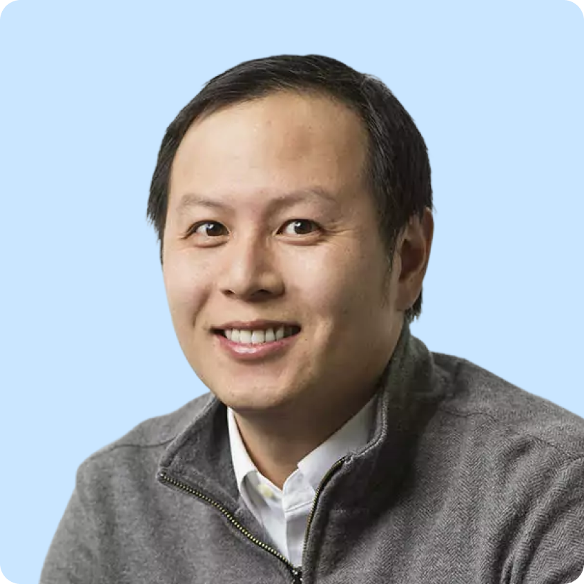 Smiling man wearing a gray zip-up sweater and white shirt against a light purple background.