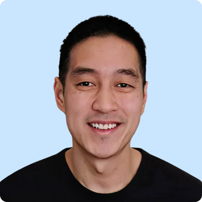 Smiling young man with short black hair wearing a black shirt against a light purple background.
