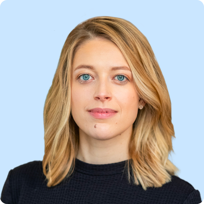 Portrait of a blonde woman with blue eyes wearing a black top against a light purple background.