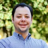 Smiling man with short black hair wearing a blue checkered shirt in front of green foliage.