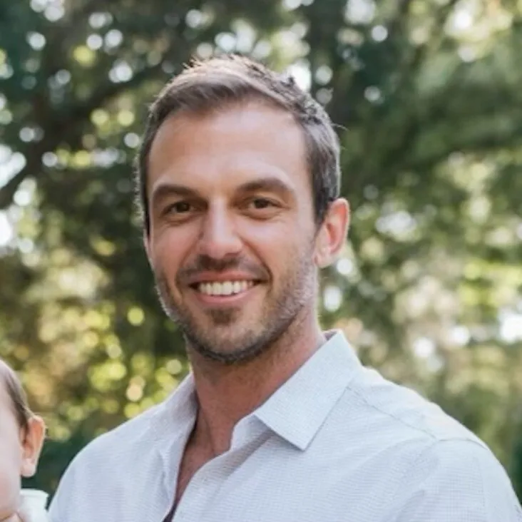 Man with short brown hair and light stubble wearing a white collared shirt smiling outdoors with trees in the background.