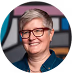 Smiling woman with short gray hair and glasses wearing a dark top in front of a colorful blurred background.