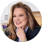 Smiling woman with long blonde hair wearing a dark blazer in a bright indoor setting.