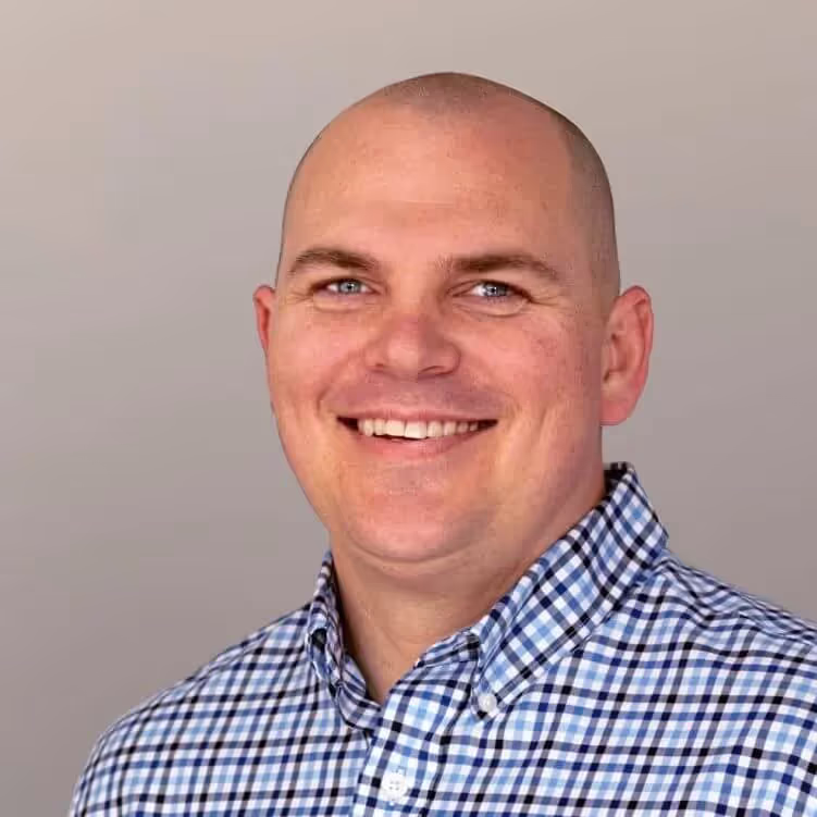 Smiling bald man wearing a blue and white checkered shirt against a light gray background.