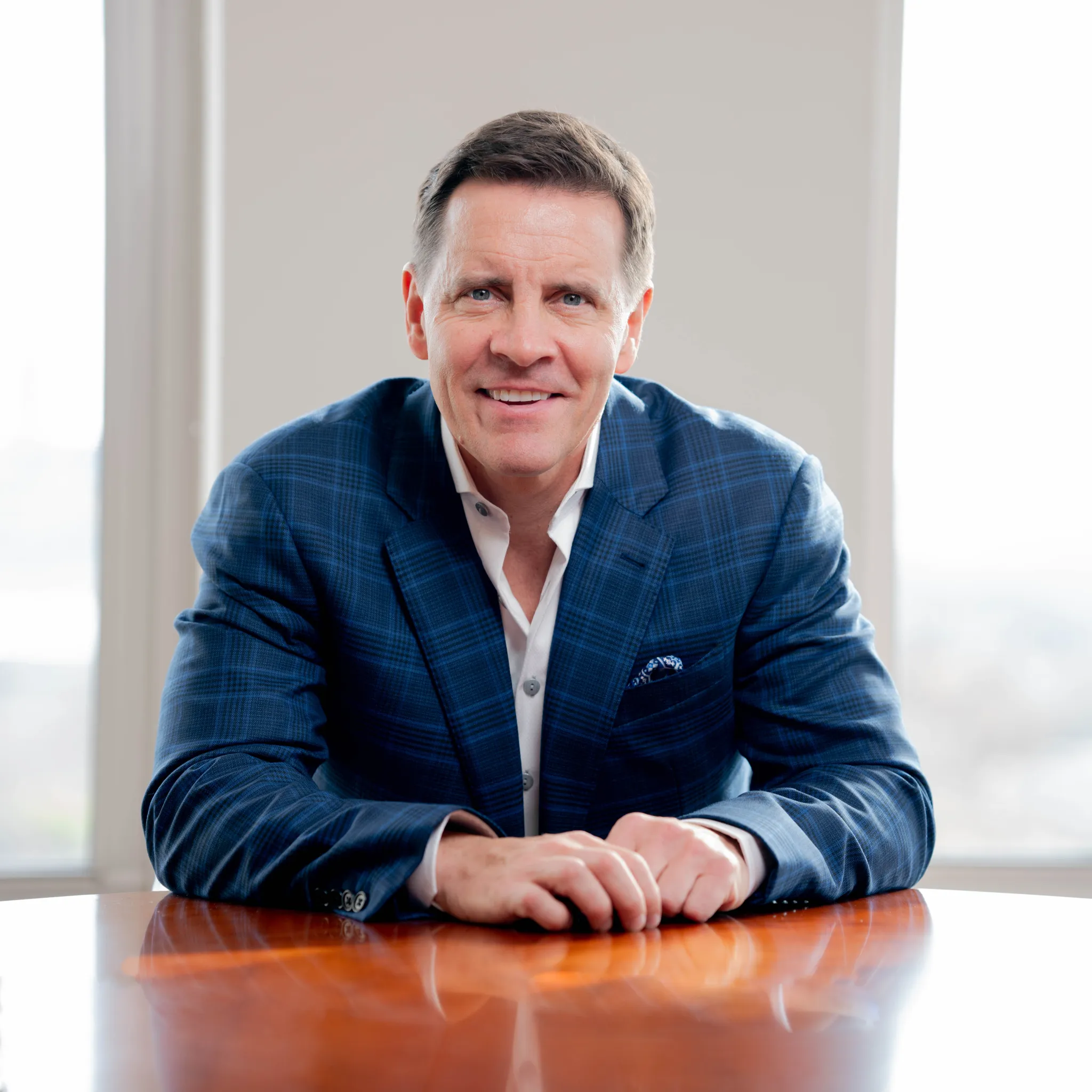 Smiling middle-aged man in a blue checkered blazer sitting at a wooden table with hands folded.