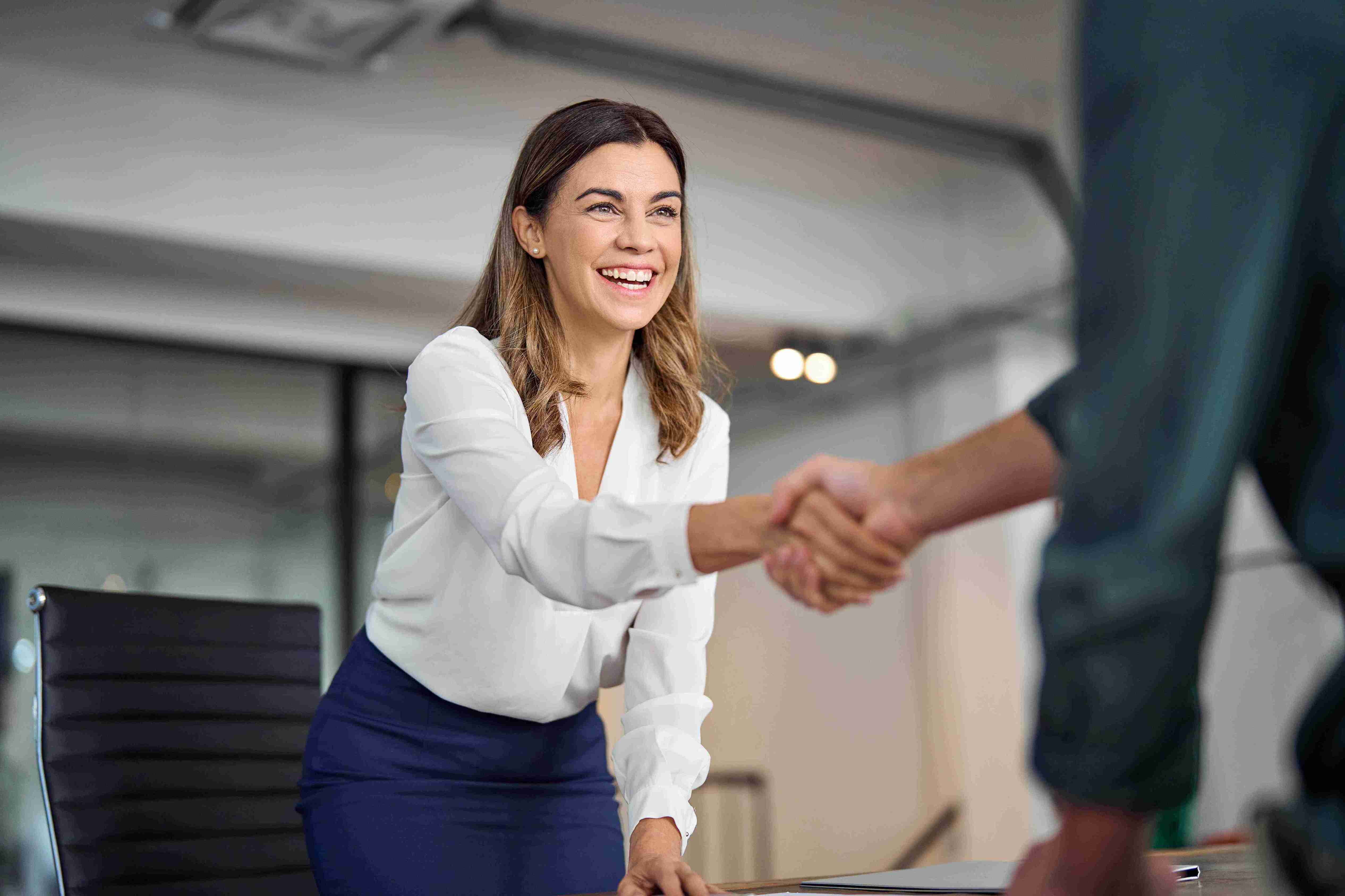 A businesswoman reaches across a desk for a handshake with an offscreen man, smiling.