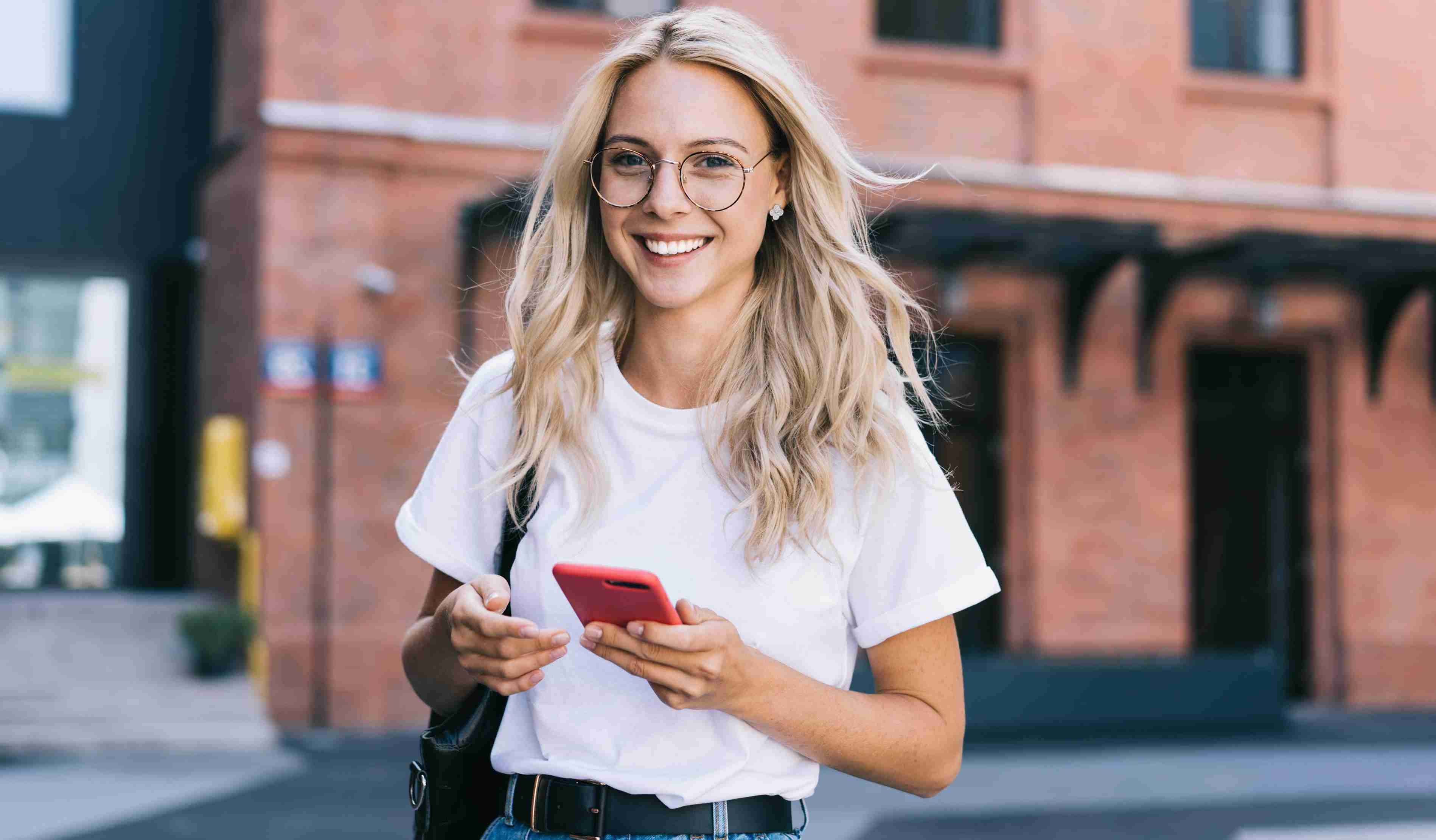 A blonde woman stands on the street outside an apartment building, smiling to the camera as she holds a phone in a red case.