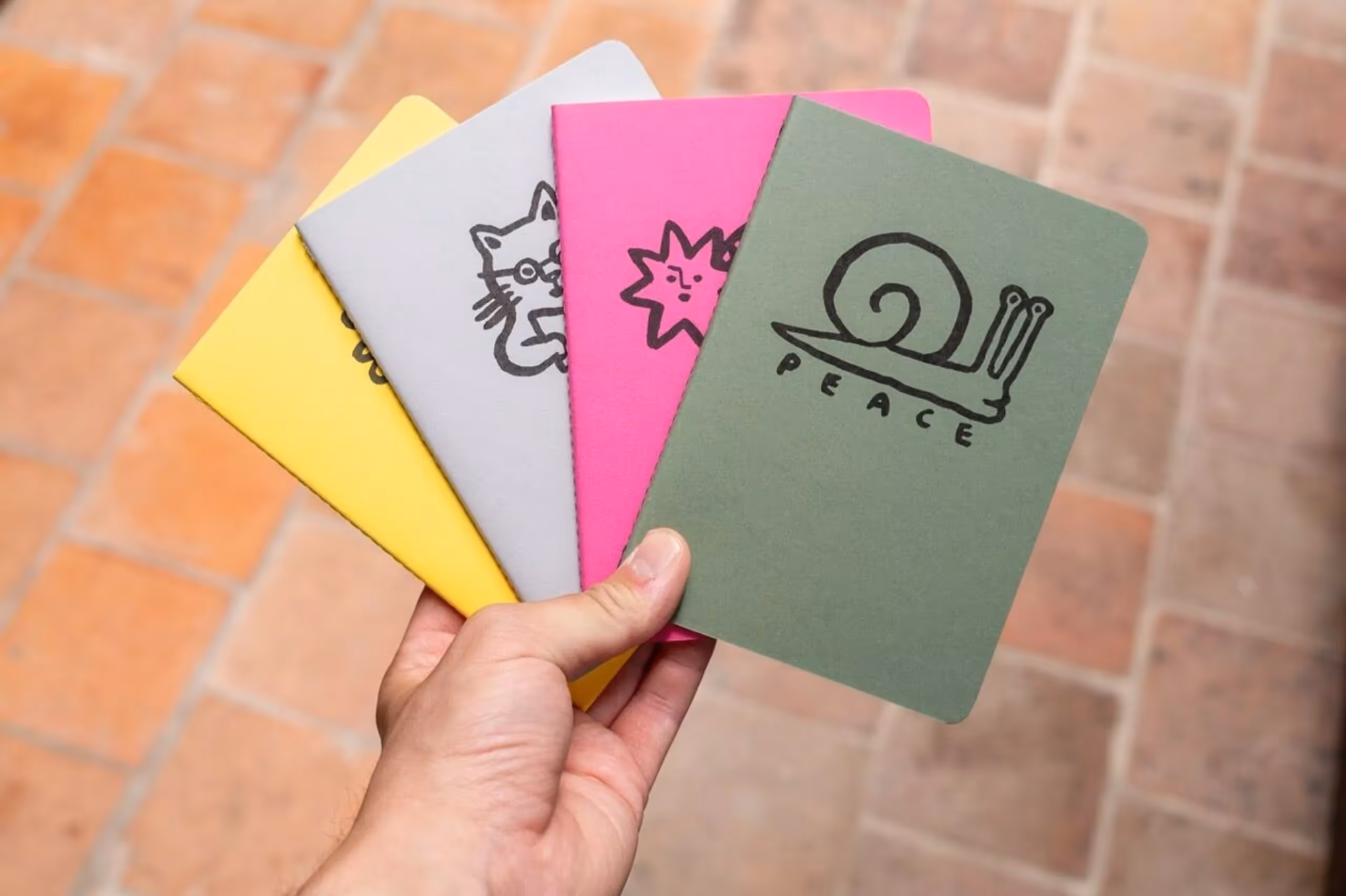 Main tenant une série de quatre carnets colorés avec des illustrations dessinées, dont un escargot avec le mot paix.
