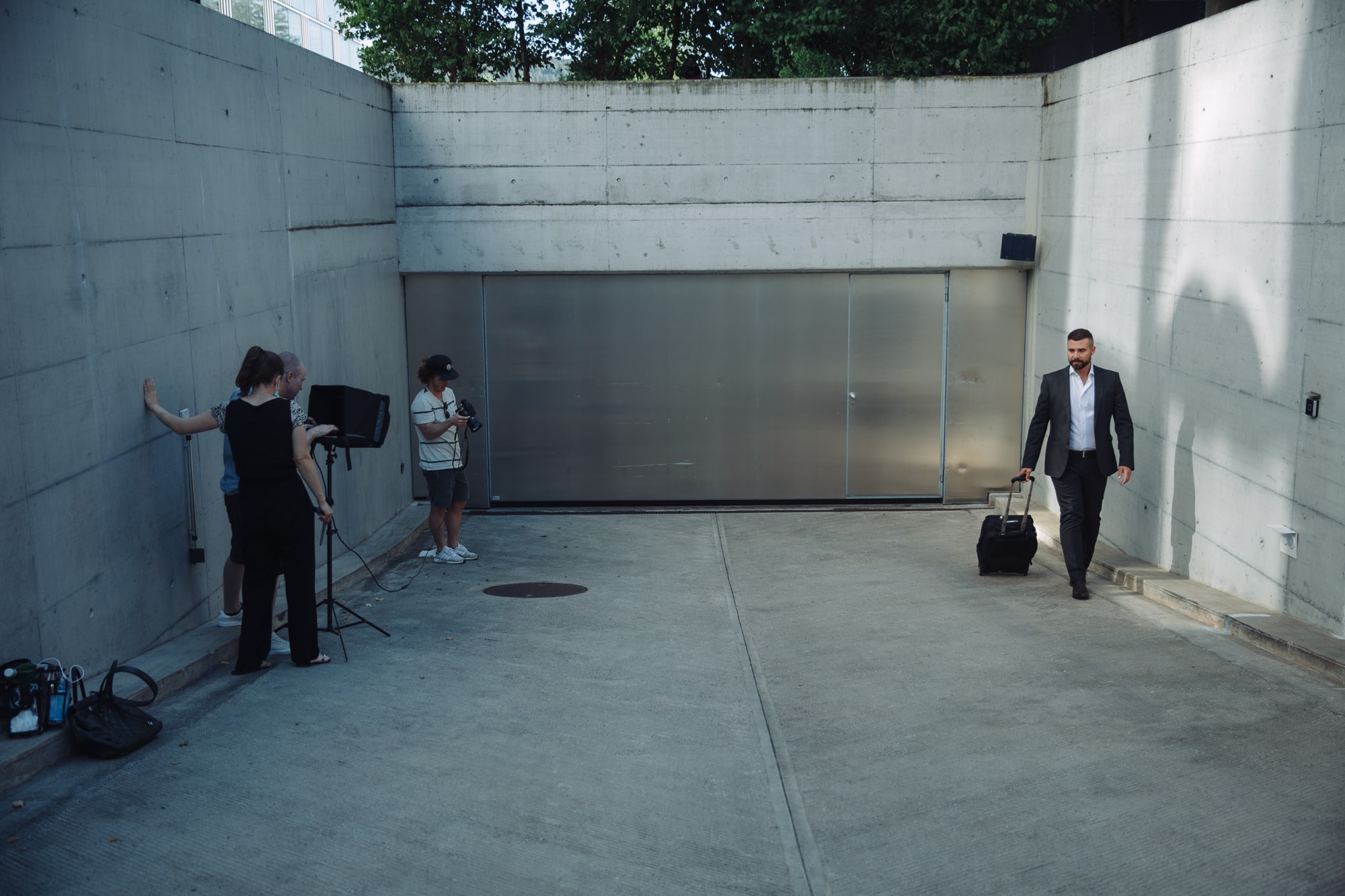 Actor in suit walking on concrete set with film crew and luggage nearby.