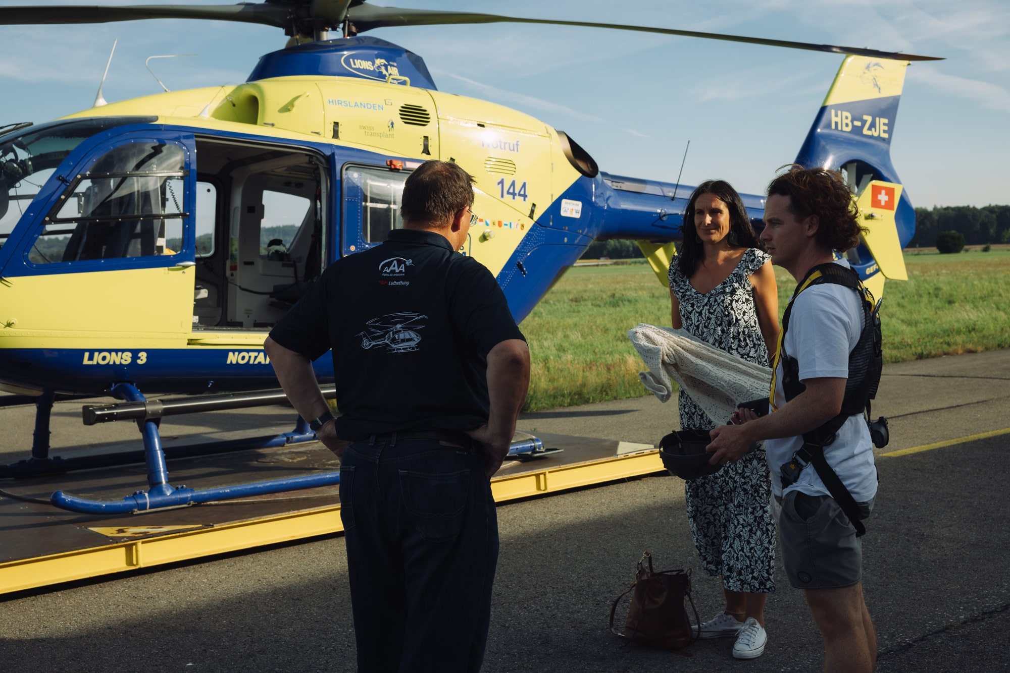 People standing beside a yellow and blue helicopter with open door on airport tarmac.