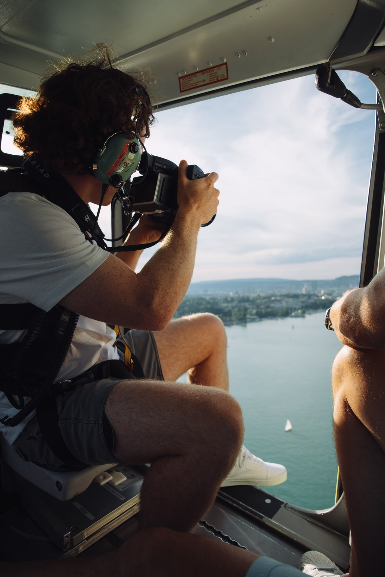 Man taking aerial photos from a helicopter overlooking a scenic urban waterfront landscape.