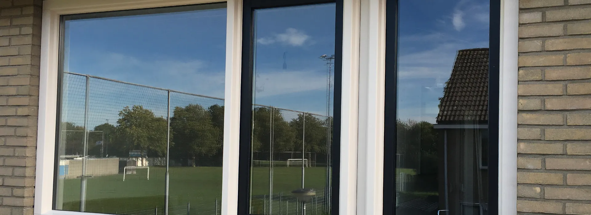Woningraam met wit kunststof kozijn en zwarte tussenstijl in een licht bakstenen gevel, met reflectie van een sportveld.